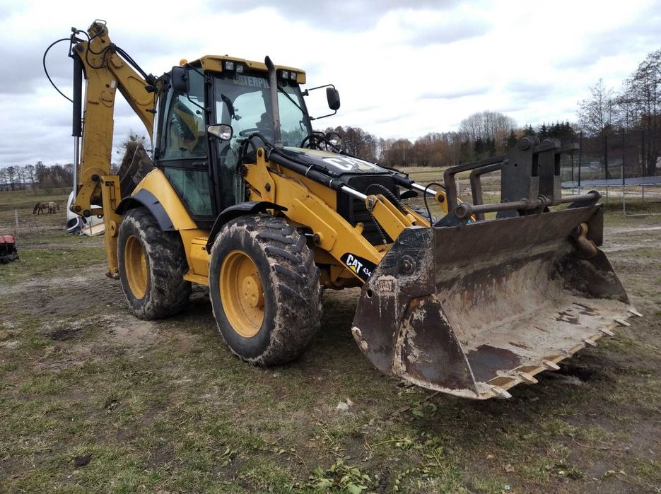 Żółta koparko-ładowarka Caterpillar 434 stoi na trawiastym terenie, z widocznymi śladami użytkowania na łyżce i oponach. W tle pastwisko z końmi pod pochmurnym niebem.