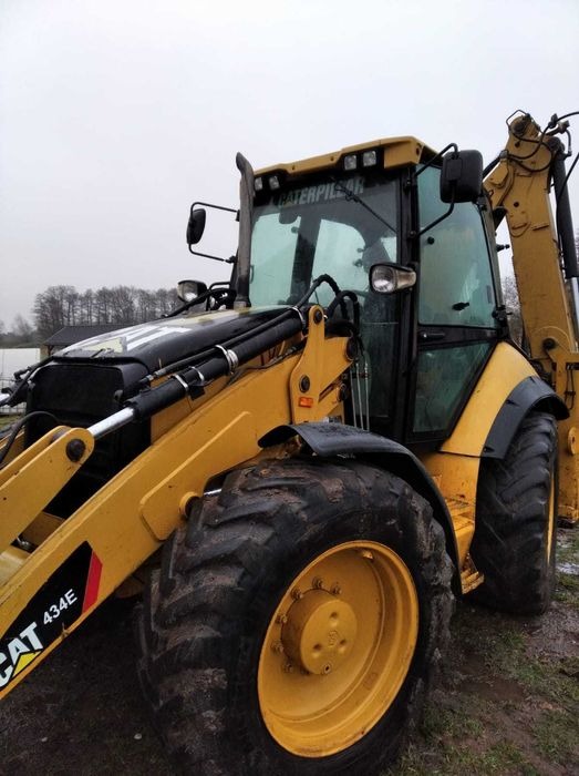 Żółta koparko-ładowarka Caterpillar 434E na wilgotnym terenie, widok z przodu, z widocznymi oponami z bieżnikiem i elementami hydraulicznymi.