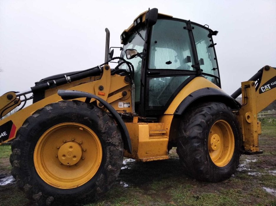 Żółta koparko-ładowarka CAT 434E z widocznymi śladami błota na oponach i karoserii, stojąca na grząskim terenie w pochmurny dzień.