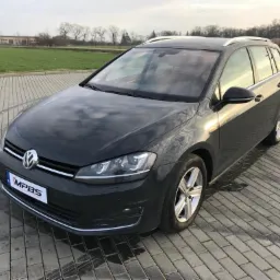 Czarny samochód kombi marki Volkswagen Golf zaparkowany na brukowanym placu, widoczne pole w tle.