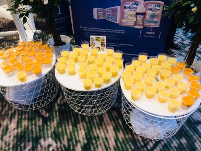 Trzy białe, druciane stoliki z małymi, plastikowymi kubeczkami wypełnionymi sokiem pomarańczowym i żółtym, ustawione na wzorzystej wykładzinie, z reklamą Bebilon w tle.