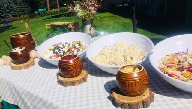 Stół z przekąskami na zewnątrz: białe miski z sałatkami, drewniane beczułki z sosami na plastrach drewna, kwiaty w tle, krata obrus