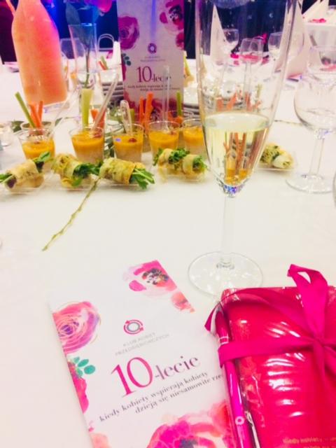 Stół z przekąskami w małych naczyniach, kieliszek z białym winem, folder reklamowy i prezent zapakowany w różowy papier z kokardą, ustawione na białym obrusie.