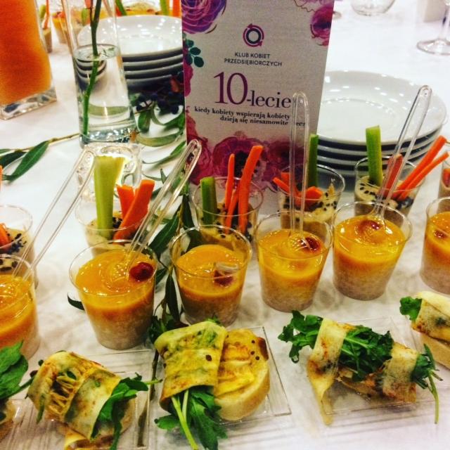 Elegancko podane przekąski: roladki z warzywami na pieczywie i mini-desery w przezroczystych kubeczkach z warzywnymi dekoracjami, całość na białym stole z dekoracją kwiatową i kartą z okazji...