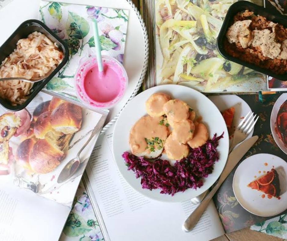 Kompozycja dań na magazynie kulinarnym: kluski ziemniaczane z sosem, czerwona kapusta, sałatka w pojemniku, różowy koktajl i ciasto pomarańczowe na talerzyku.