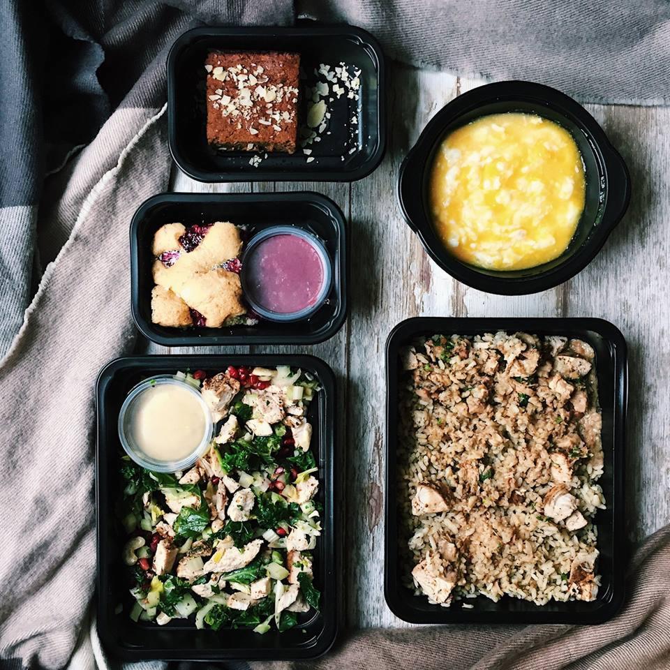 Zestaw pięciu dań dietetycznych w czarnych pojemnikach: brownie z migdałami, ciasteczka z dżemem i sosem, zupa krem, sałatka z kurczakiem i granatem oraz ryż z kurczakiem, ułożone na drewnianym...