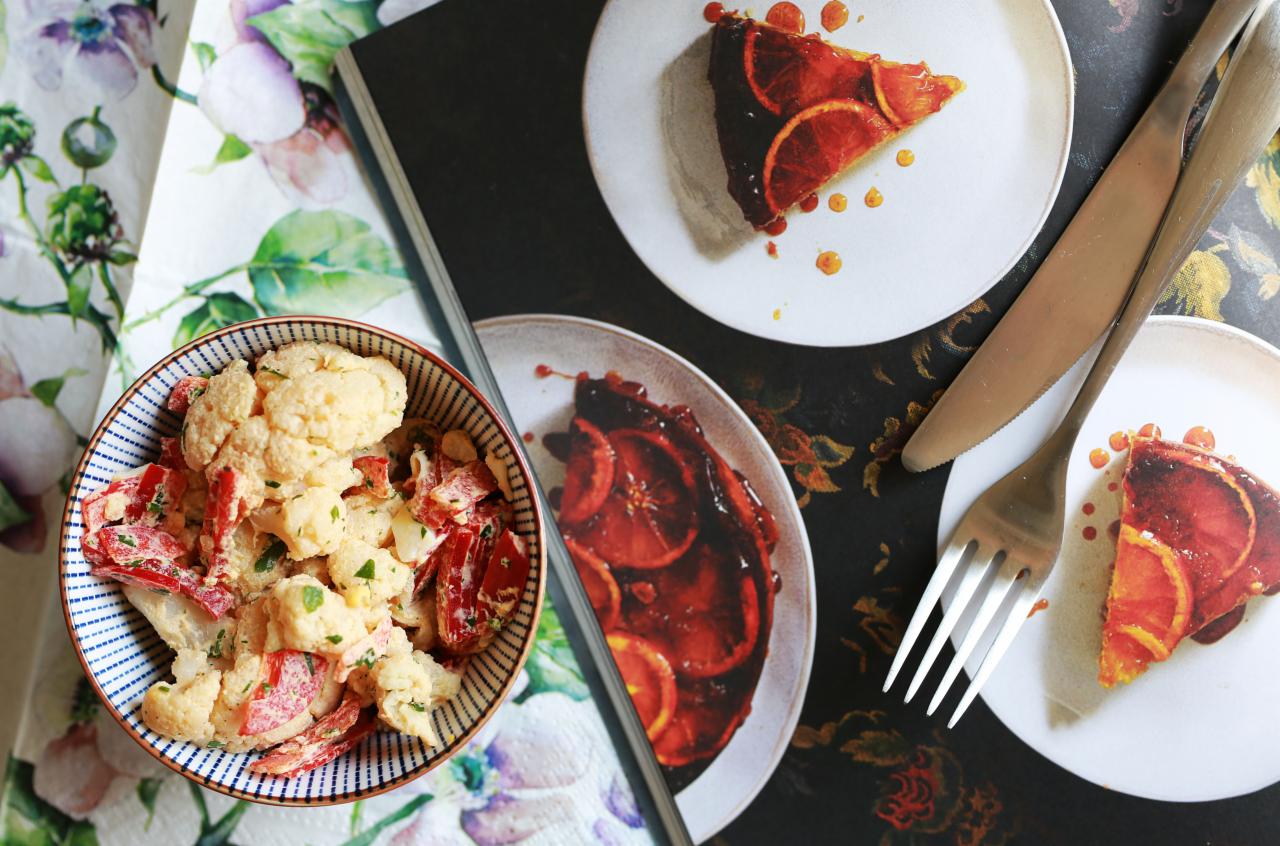Kompozycja z sałatką z kalafiora i papryki w miseczce oraz zdjęciami ciasta pomarańczowego na talerzykach, nóż i widelec leżą na czarnym tle z kwiatowym wzorem.