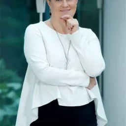 Portret kobiety w białej bluzce i czarnej spódnicy, z ręką podpartą pod brodą, sugerujący rozważania lub planowanie, na neutralnym tle.