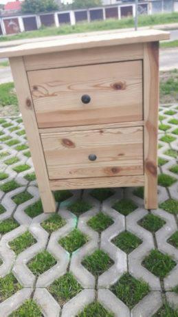 Surowa sosnowa szafka nocna z dwoma szufladami, wykończona czarnymi okrągłymi gałkami, ustawiona na betonowej ażurowej nawierzchni z zieloną trawą.