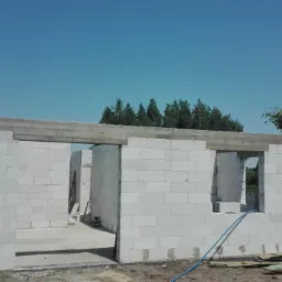 Surowy stan domu z betonu komórkowego w trakcie budowy, widoczne otwory okienne i drzwiowe, zbrojony betonowy nadproże, niebieski wąż na ziemi.