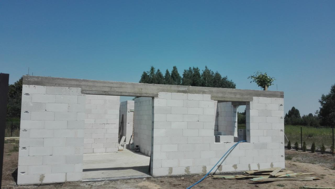 Surowy stan domu z betonu komórkowego w trakcie budowy, widoczne otwory okienne i drzwiowe, zbrojony betonowy nadproże, niebieski wąż na ziemi.