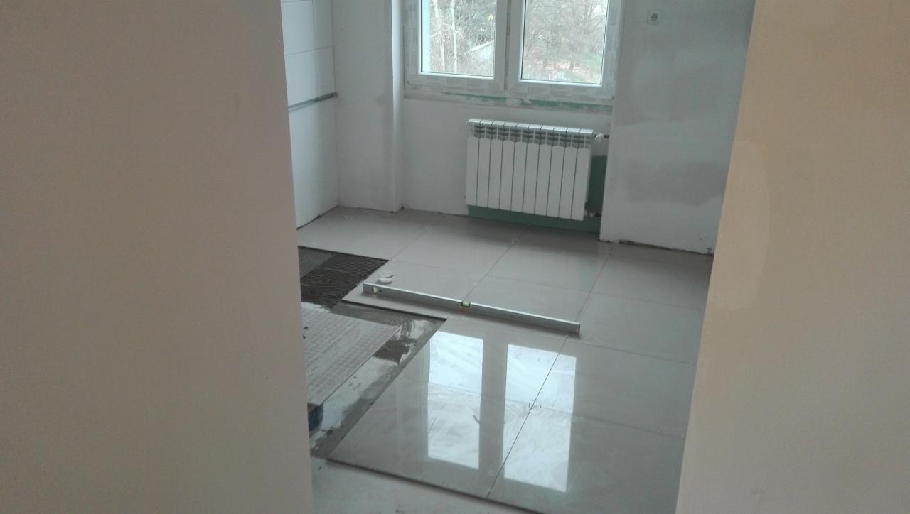 Układanie jasnych, błyszczących płytek podłogowych w pomieszczeniu z grzejnikiem pod oknem, poziomica spoczywa na płytkach.