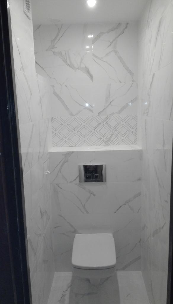 Wykończona toaleta z białymi marmurkowymi płytkami, geometrycznym wzorem nad ukrytą spłuczką i minimalistyczną muszlą klozetową.