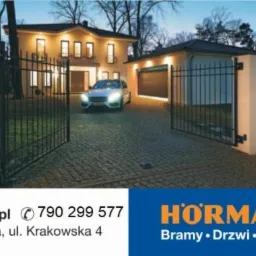 Otwarta, czarna, metalowa brama wjazdowa z dwoma słupkami, prowadząca na posesję z domem i garażem; na podjeździe stoi srebrny samochód z włączonymi światłami.