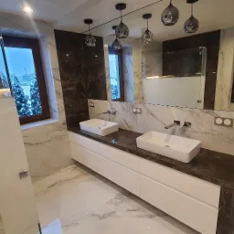 Nowoczesna łazienka z marmurowymi ścianami, podwójną umywalką nablatową na białej szafce z ciemnym blatem, szklaną kabiną prysznicową i wiszącymi lampami w kształcie kul.