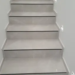 Schody wyłożone jasnymi płytkami z czarnymi paskami antypoślizgowymi, oświetlone kwadratowymi lampami ściennymi LED, widok z dołu.