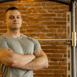 Mężczyzna z założonymi rękami na tle ściany z cegły i sprzętu do ćwiczeń.