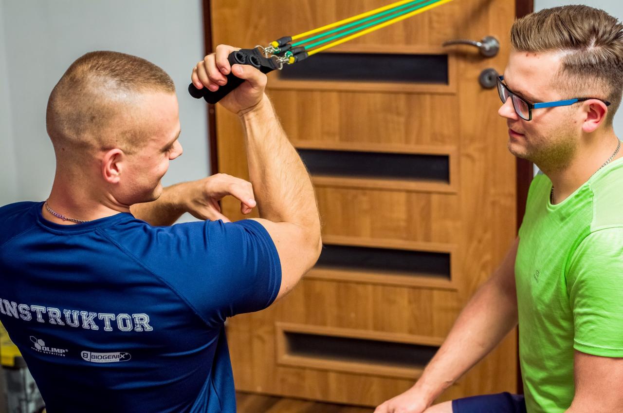 Mężczyzna w niebieskiej koszulce z napisem 'INSTRUKTOR' ćwiczy biceps z gumami oporowymi, drugi mężczyzna w okularach obserwuje.