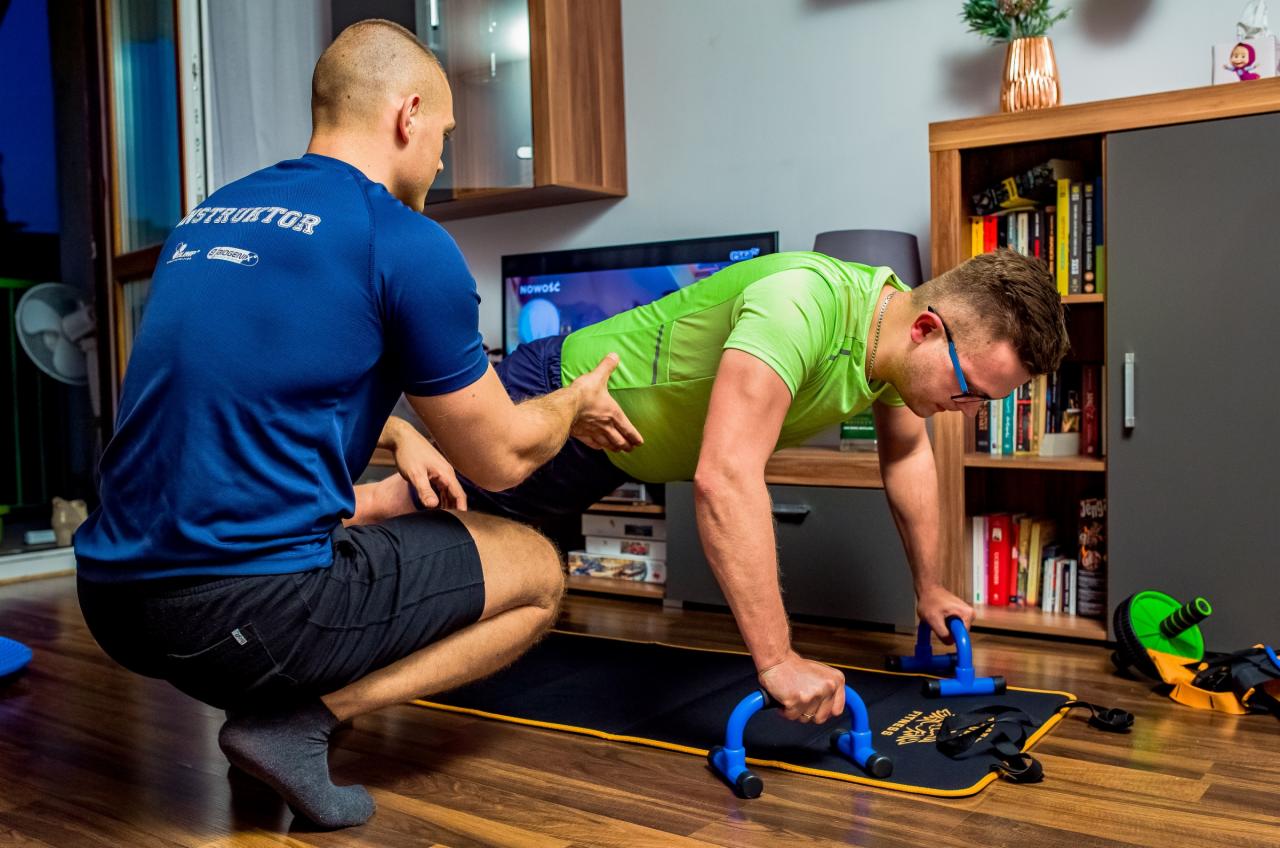 Mężczyzna w zielonej koszulce ćwiczy pompki na uchwytach, instruktor w niebieskiej koszulce asekuruje go, w tle widoczny regał z książkami i telewizor.