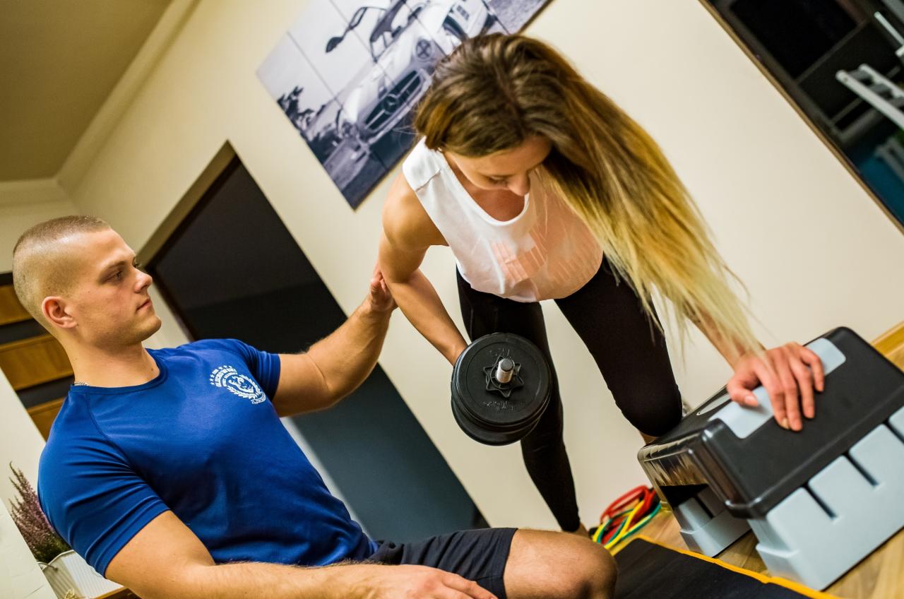 Kobieta ćwiczy z hantlą, opierając się o step, pod nadzorem mężczyzny w niebieskiej koszulce z logo, który asekuruje jej ramię, w tle plakat z samochodem.