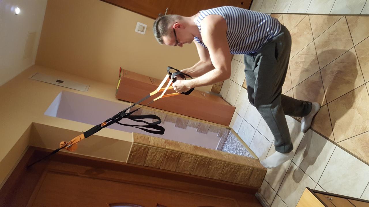 Mężczyzna w okularach i paski koszulce ćwiczy z zawieszonymi pasami do treningu TRX w pomieszczeniu o beżowych ścianach i kafelkach.