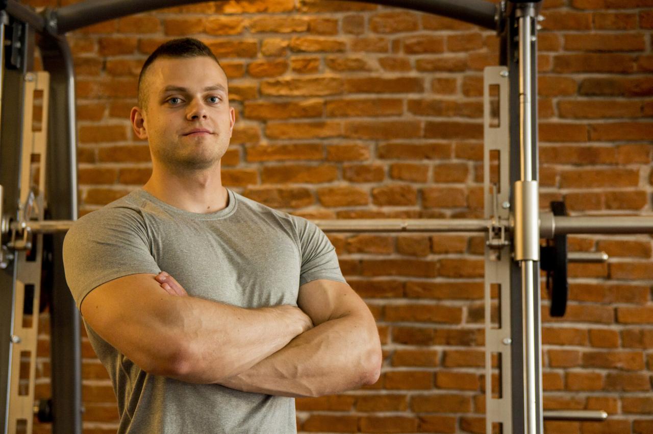 Mężczyzna z założonymi rękami na tle ściany z cegły i sprzętu do ćwiczeń.