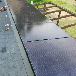 Częściowy widok instalacji paneli słonecznych na dachu pokrytym gontem bitumicznym. Widoczne narzędzia i elementy konstrukcyjne w tle. Wysoki kąt ujęcia.