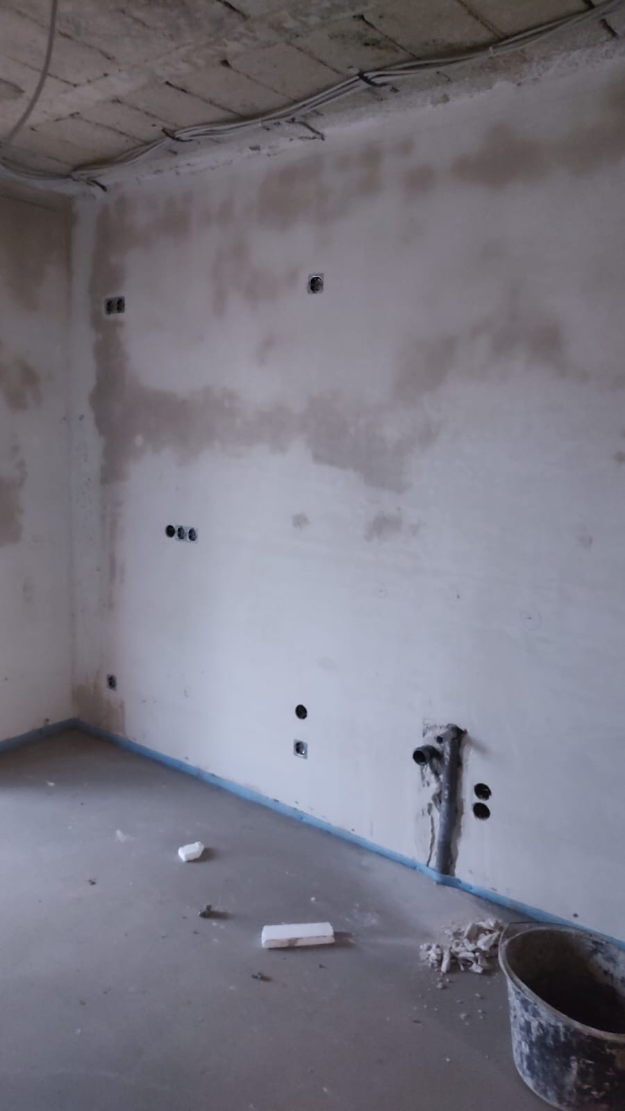 Surowy stan wnętrza: ściany po wstępnym tynkowaniu z widocznymi puszkami elektrycznymi i wyprowadzonymi przewodami. Podłoga betonowa, widoczne kable pod sufitem.