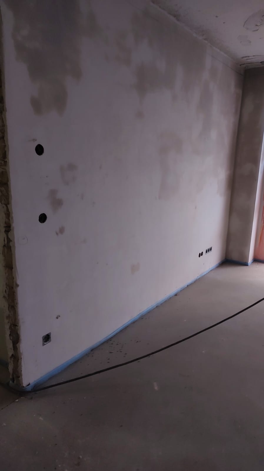 Ściana po tynkowaniu z widocznymi otworami pod gniazdka elektryczne i kable na podłodze, przygotowana do dalszych prac wykończeniowych.