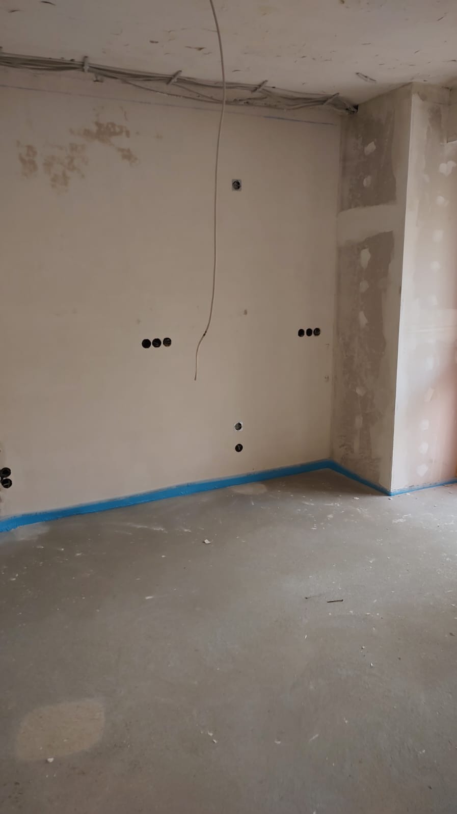 Surowy stan wnętrza z instalacją elektryczną: kable pod sufitem, otwory na gniazdka w ścianie, zabezpieczona podłoga taśmą. Widok na przygotowane ściany i surową podłogę.