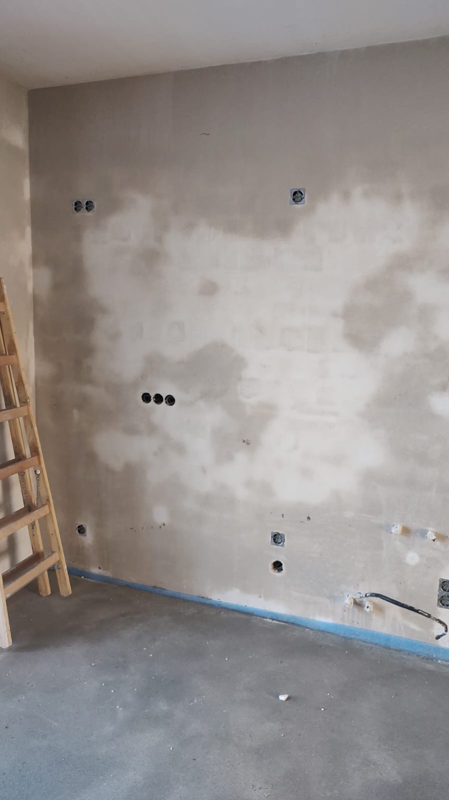 Ściana w trakcie remontu z widocznymi puszkami elektrycznymi i wystającymi przewodami, drabina drewniana oparta o ścianę, surowy beton na podłodze.