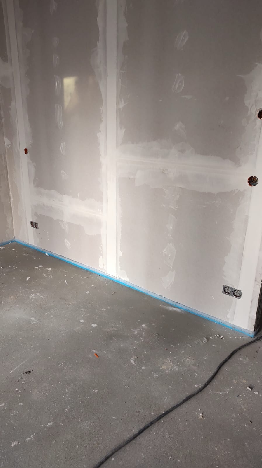 Ściana w trakcie szpachlowania z widocznymi puszkami elektrycznymi i zabezpieczeniem podłogi niebieską folią. W tle widoczny kabel elektryczny.