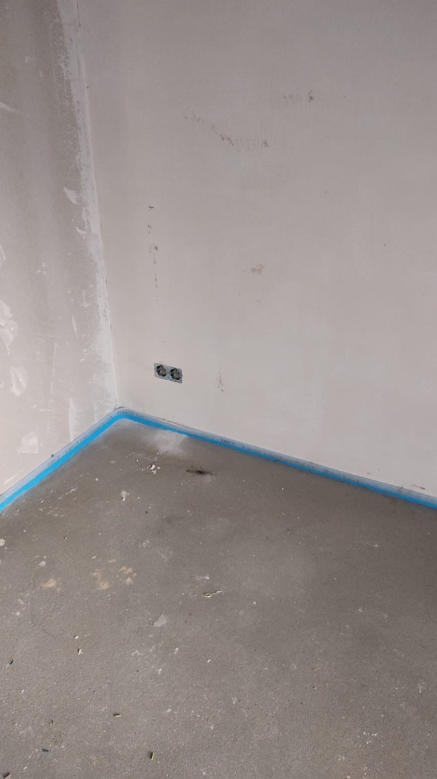Surowy stan: gładka wylewka podłogowa zabezpieczona niebieską taśmą, puszki elektryczne w ścianie, przygotowanie do dalszych prac wykończeniowych.