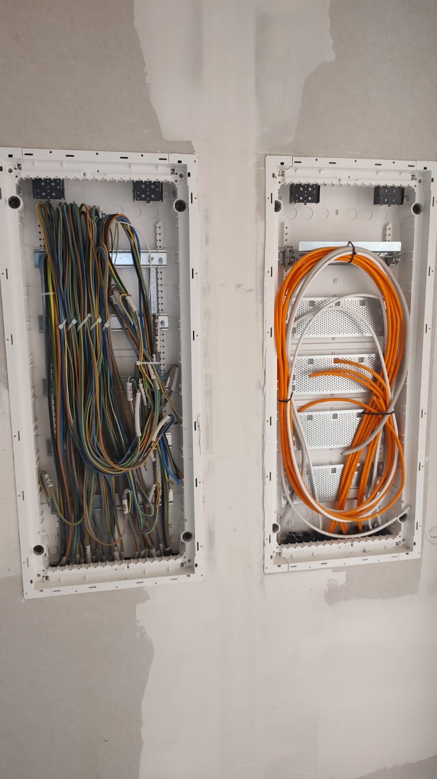Dwa białe panele z przewodami: kolorowe po lewej, pomarańczowe po prawej, zamontowane na surowej ścianie w trakcie instalacji elektrycznej.