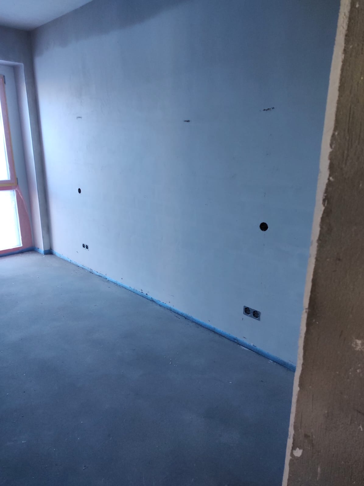 Wnętrze w trakcie prac wykończeniowych: gładkie ściany z widocznymi puszkami instalacyjnymi i gniazdkami, zabezpieczona podłoga, surowy stan deweloperski.