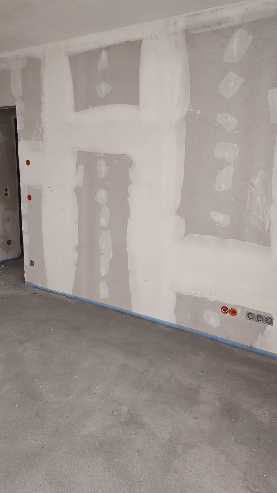 Wnętrze w trakcie prac wykończeniowych: ściany z widocznymi punktami elektrycznymi, częściowo wyszpachlowane, surowa podłoga, zabezpieczona taśmą.