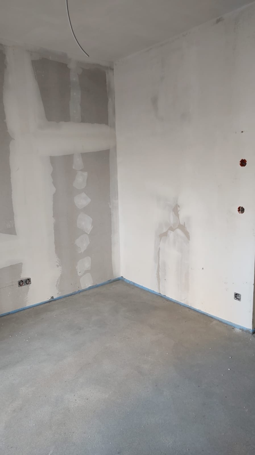 Surowy stan wnętrza: ściany po gruntowaniu, widoczne puszki elektryczne, wylewka podłogowa zabezpieczona taśmą. Kabel zwisa z sufitu.