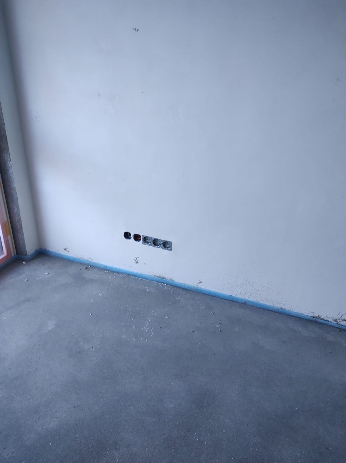 Surowa ściana z zamontowanymi gniazdkami elektrycznymi i otworem. Podłoga betonowa z niebieską taśmą zabezpieczającą przy ścianie. Wnętrze w trakcie prac wykończeniowych.