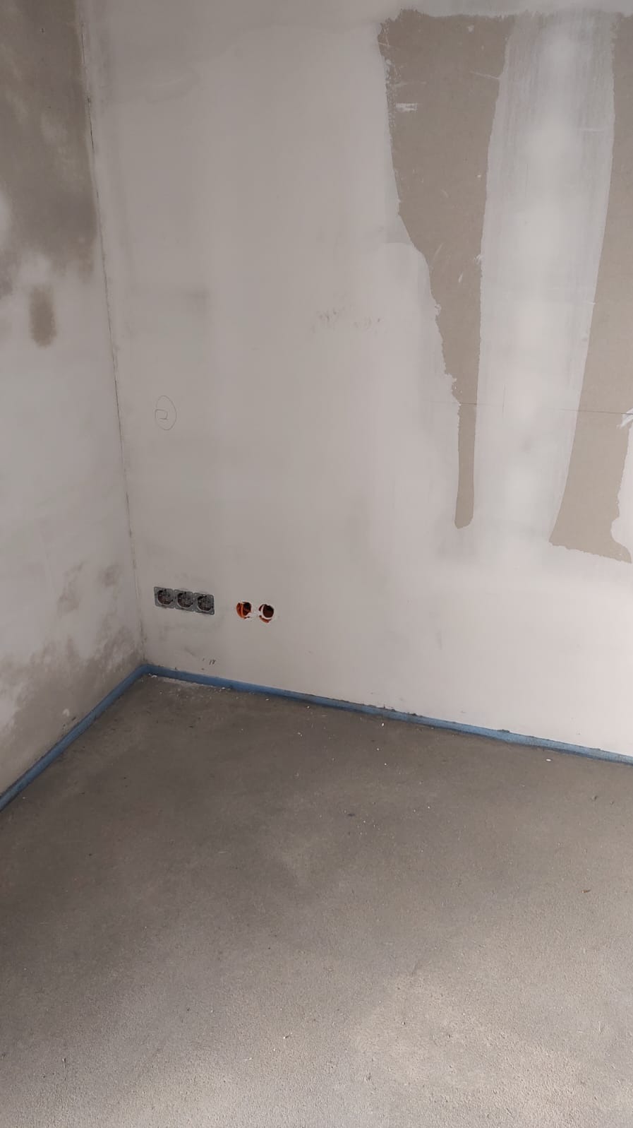 Surowy stan wnętrza z widocznymi gniazdkami elektrycznymi i instalacją w ścianie, zabezpieczona podłoga folią w narożniku, ściany częściowo wykończone gładzią.