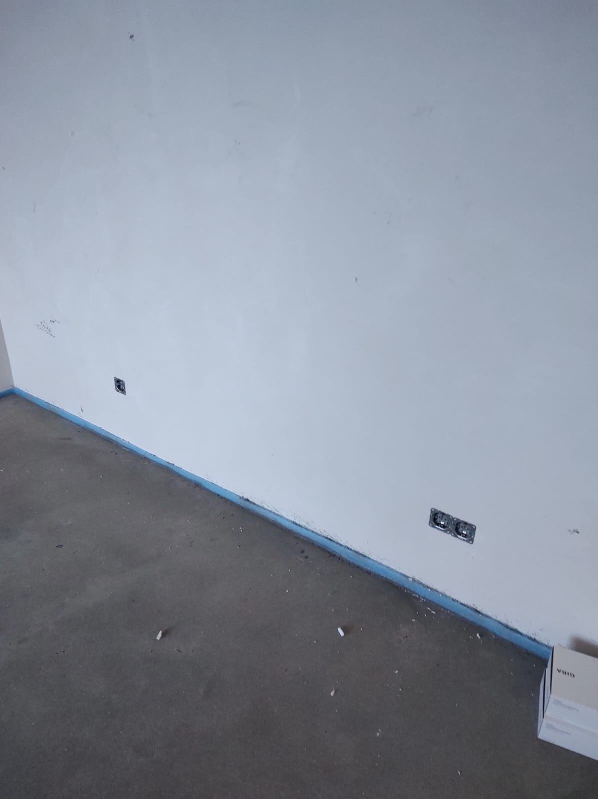 Surowa ściana z puszkami elektrycznymi, zabezpieczona niebieską taśmą malarską przy podłodze. Widoczna skrzynka GIRA. Minimalizm w trakcie prac wykończeniowych.