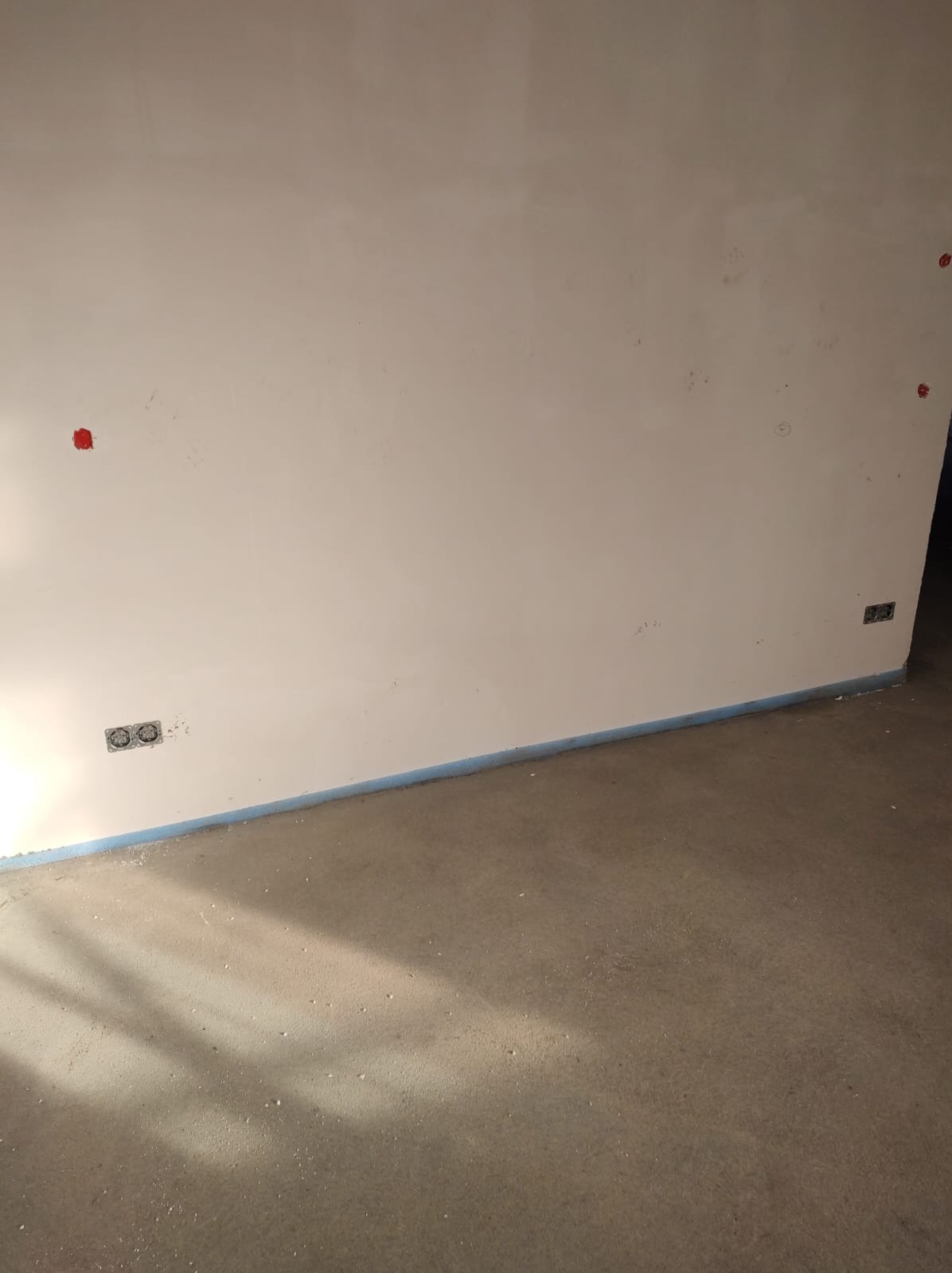 Ściana z widocznymi otworami na gniazdka elektryczne zabezpieczonymi czerwonymi zaślepkami, oraz wykończona podłoga z listwą przypodłogową zabezpieczoną taśmą.