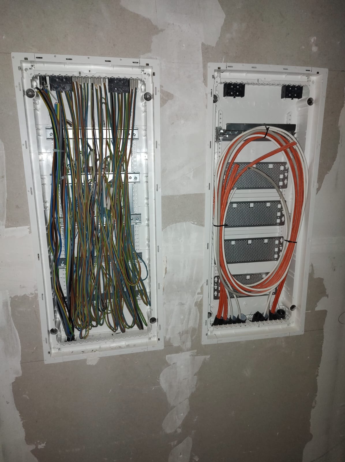 Dwa panele elektryczne w ścianie z przewodami: jeden z wieloma kolorowymi kablami, drugi z pomarańczowymi i białymi kablami ułożonymi w pętle. Szara ściana w tle.