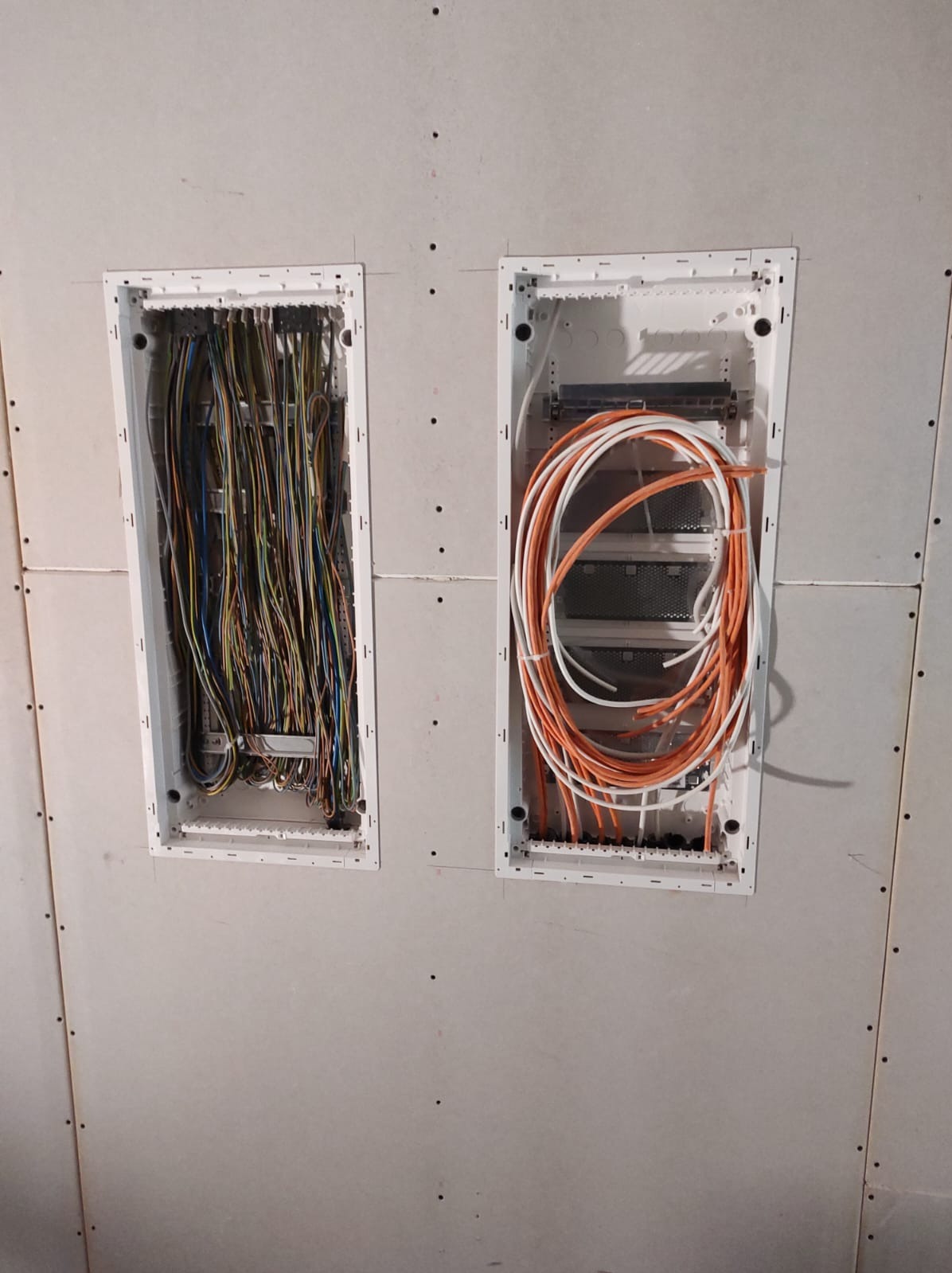 Dwa otwory w ścianie z płyt gipsowych, wewnątrz widoczne kable elektryczne: kolorowe i pomarańczowo-białe. Instalacja w trakcie prac, przed wykończeniem.