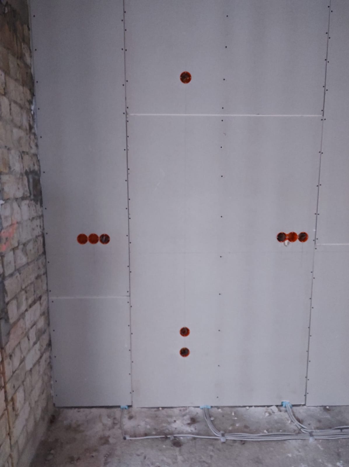 Ściana z płyt gipsowo-kartonowych z widocznymi puszkami instalacyjnymi pod gniazdka elektryczne i kable na podłodze. Surowy stan wykończenia wnętrza.
