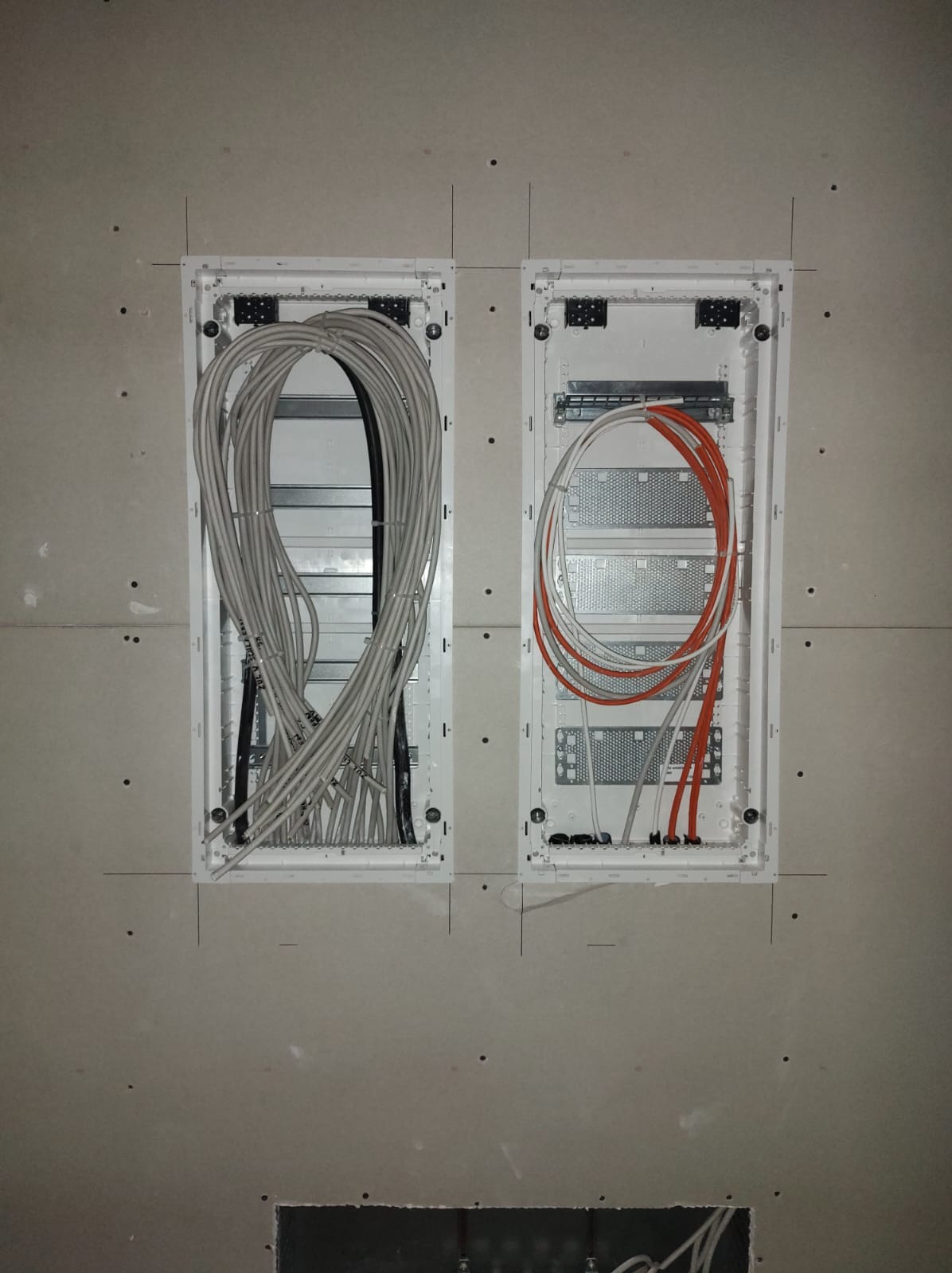 Dwie białe skrzynki elektryczne w ścianie z płyt gipsowych. Lewa pełna kabli szarych i czarnych, prawa z przewodami pomarańczowymi i szarymi. Widoczne oznaczenia na kablach.