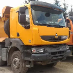 Dwa samochody ciężarowe wywrotki: żółty Renault Kerax z plandeką i pomarańczowy Mercedes-Benz, zaparkowane na placu budowy.