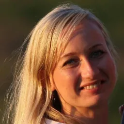 Portret uśmiechniętej blondynki z niebieskimi oczami, oświetlonej ciepłym światłem, na rozmytym, zielonym tle. Widoczne zmarszczki mimiczne.