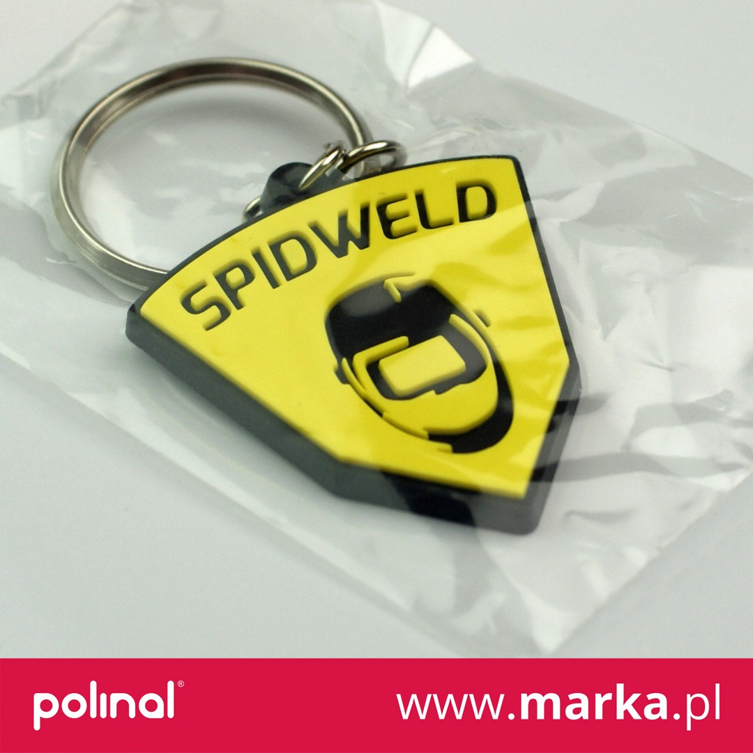 Żółto-czarny brelok w kształcie tarczy z logo firmy SPIDWELD i symbolem maski spawalniczej, zawieszony na metalowym kółku, zapakowany w przezroczysty woreczek.