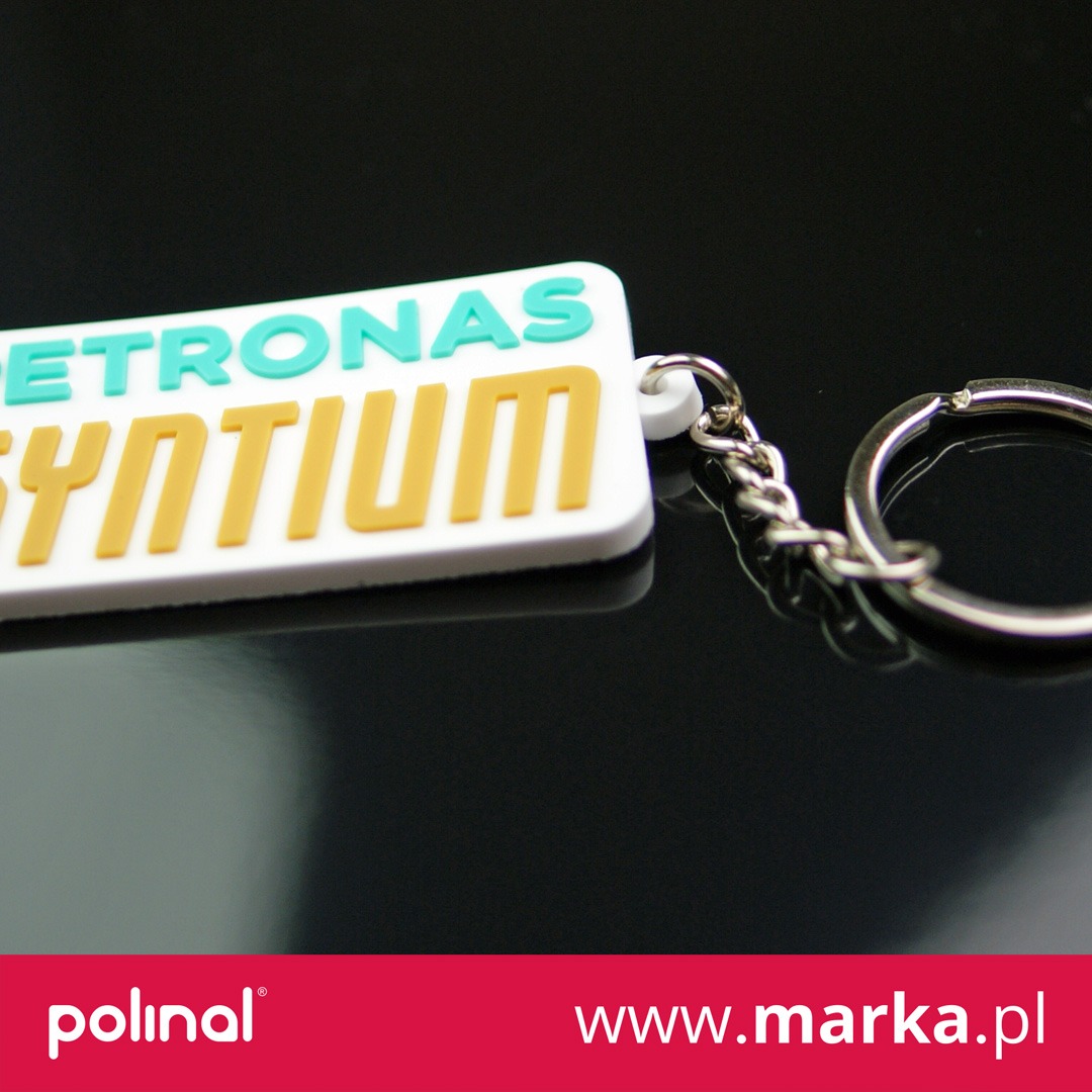 Biały brelok z logo PETRONAS Syntium w kolorach turkusowym i żółtym, zawieszony na metalowym łańcuszku, leżący na czarnym, błyszczącym tle.