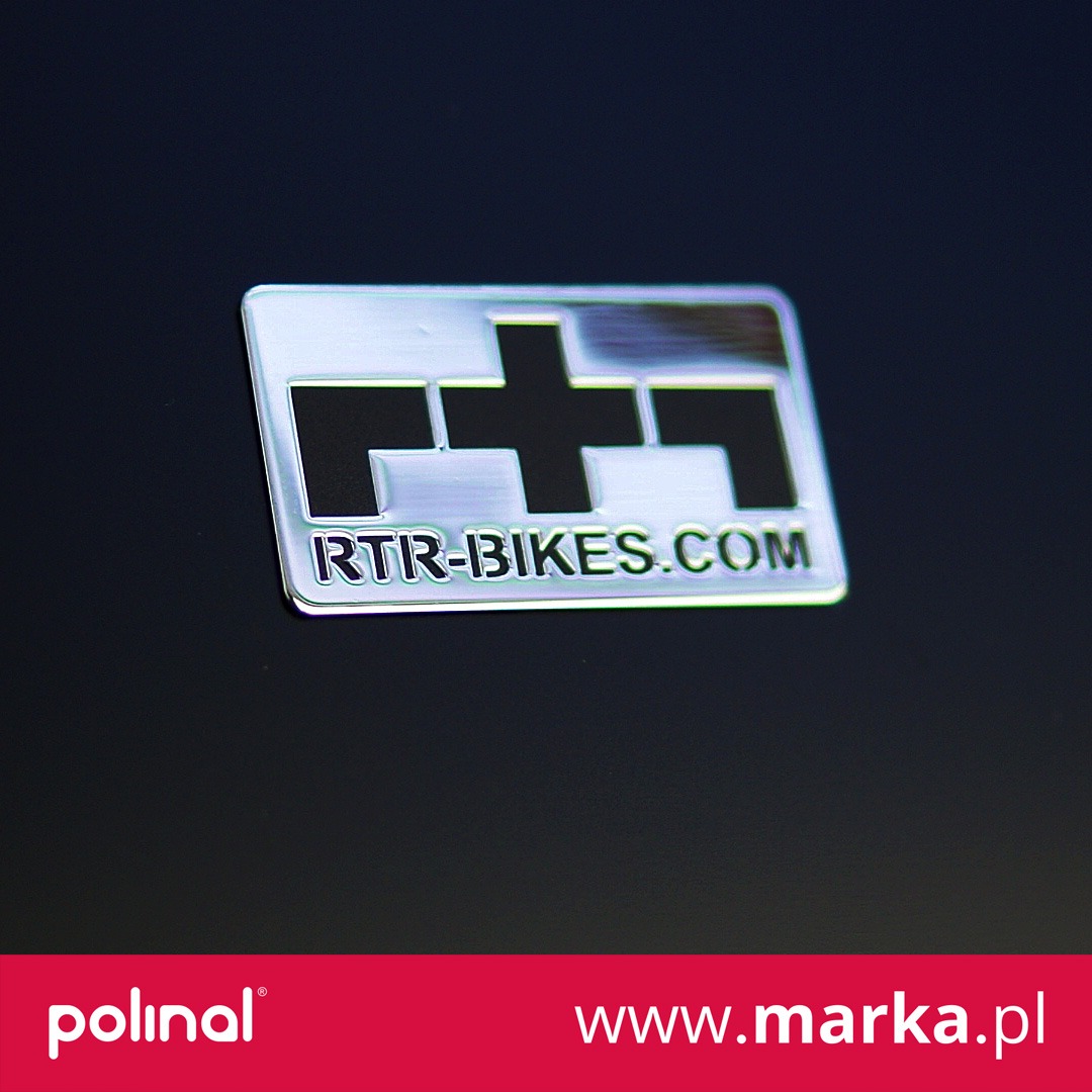 Błyszcząca, srebrna etykieta z logo RTR-BIKES.COM na ciemnym tle.
